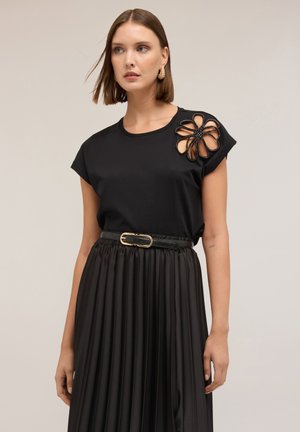 Top nero a maniche corte con un design a fiori ritagliato e decorazioni nere. Abbinato a una gonna nera plissettata e una cintura con fibbia dorata.