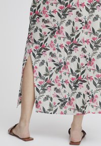 Robe longue à motifs floraux en tissu léger de couleur blanche, ornée de motifs roses et verts. Fente sur le côté et sandales à glissière marron visibles au niveau de l'ourlet.