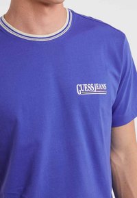 Mężczyzna w purpurowej koszulce z białym i niebieskim paskiem przy kołnierzyku oraz napisem "GUESS JEANS QUALITY" wydrukowanym na lewej piersi.