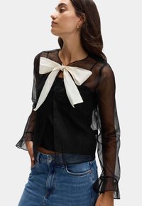 Blusa negra transparente de manga larga con un lazo blanco en el cuello. Presenta un escote redondeado y puños de manga con volantes. Jeans de mezclilla azul debajo.