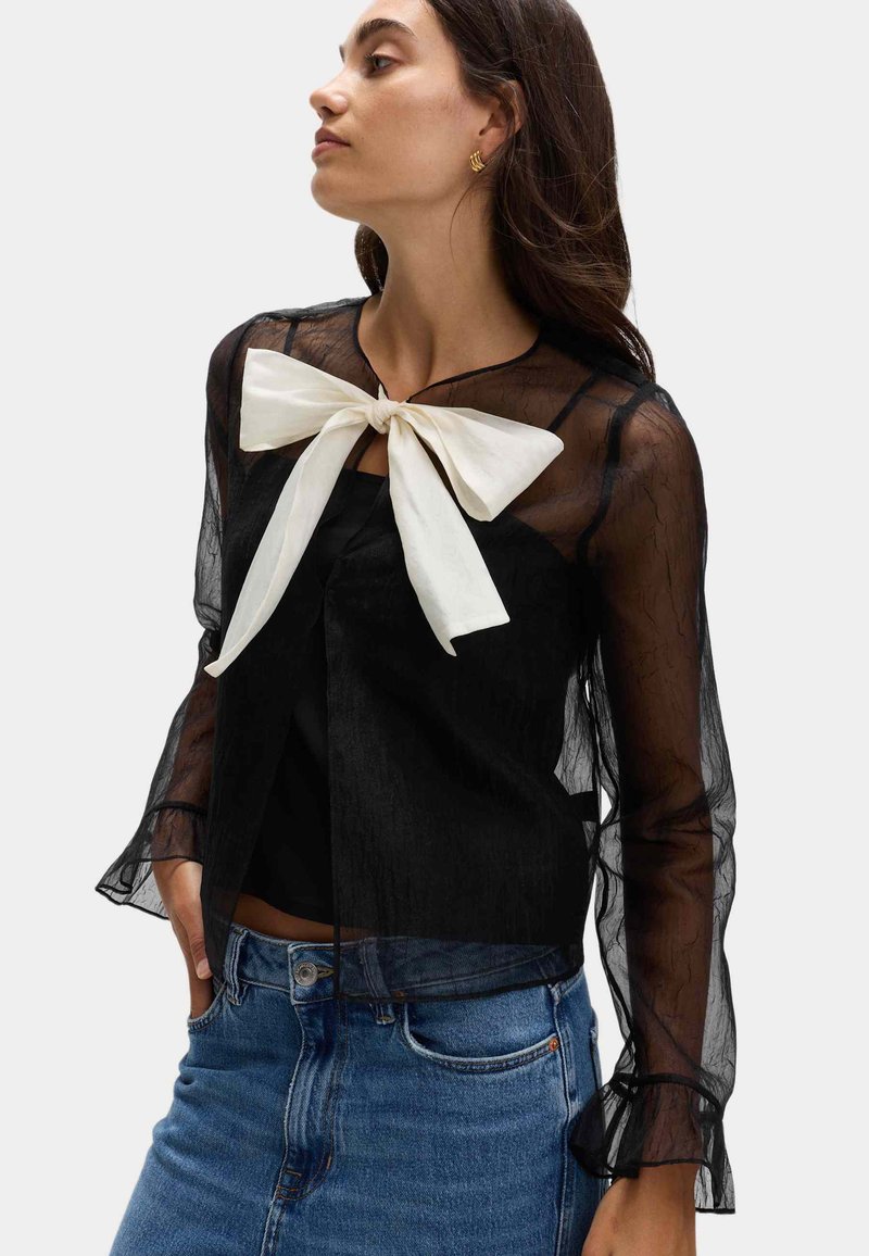Blusa negra transparente de manga larga con un lazo blanco en el cuello. Presenta un escote redondeado y puños de manga con volantes. Jeans de mezclilla azul debajo.