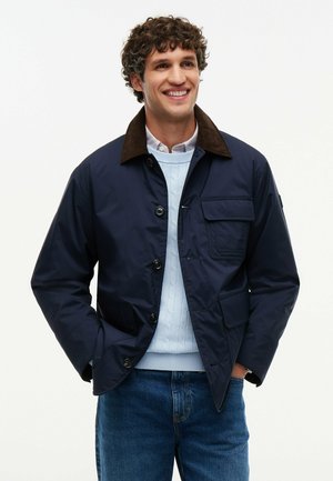 Smilende mand med krøllet hår iført en navyjakke med brun krave, lyseblå sweater og blå jeans, hænderne i lommerne.