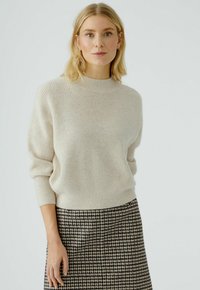 Hellbeige gerippter Pullover mit hohem Kragen und weichem Material. Über einem schwarz-weißen Hahnentrittmuster-Rock getragen. Schlichtes Design.
