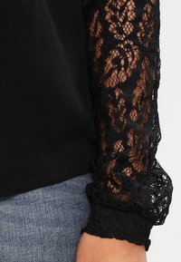 Schwarzes Oberteil mit langen, durchsichtigen Spitzenärmeln, die Blumenmuster aufweisen. Die Ärmelenden haben einen fransenartigen Saum und werden über Jeans getragen.
