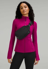 Bandolera negra llevada sobre una chaqueta rosa brillante con cremallera, que presenta mangas largas y un diseño ajustado. Leggings negros completan el atuendo.