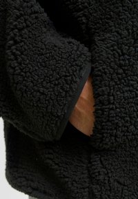 Sudadera de forro polar negro con un exterior texturizado y esponjoso. Las mangas presentan un ribete negro en los puños, dejando al descubierto una mano desnuda.