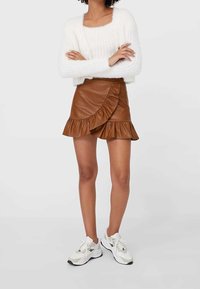 Eine Frau trägt einen weißen, flauschigen Pullover, einen braunen, fauxledernen Minirock mit Rüschen und weiße Sportschuhe, während sie mit verschränkten Armen steht.