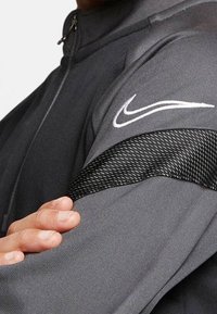 Grå idrottsjacka med en slät yta, svart vävd detalj på ärmen och en vit Nike-logotyp på axeln.