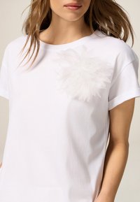 Camiseta blanca de manga corta hecha de algodón suave, con un acento floral texturizado en blanco en el lado izquierdo del pecho.