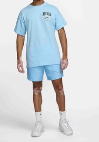 Person som bär en ljusblå Nike-t-shirt och shorts, vita Nike-strumpor och vita sneakers, står mot en enfärgad bakgrund.