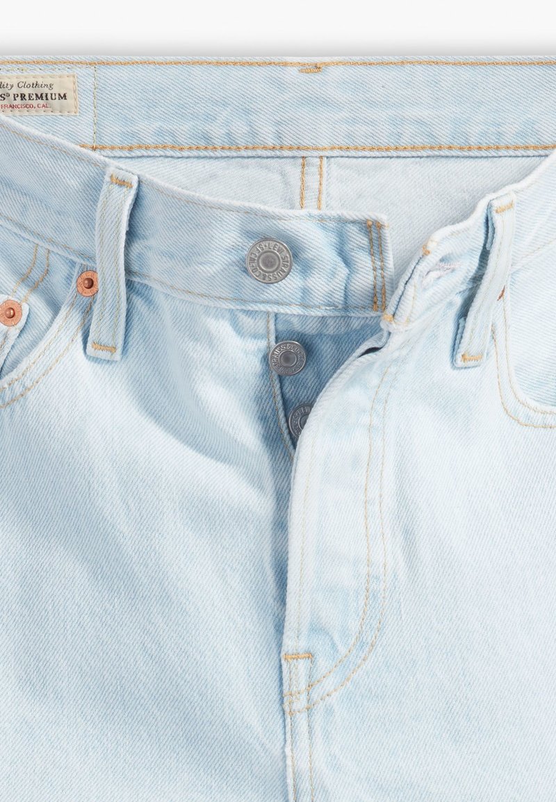 Un jean en denim bleu clair présente un design taille haute avec des coutures visibles, des rivets en cuivre et une fermeture par bouton à l'avant.