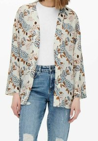 Kimono giacca floreale in tessuto crema, con fiori blu e marroni, maniche lunghe, apertura frontale, indossata su una maglietta bianca e jeans strappati.