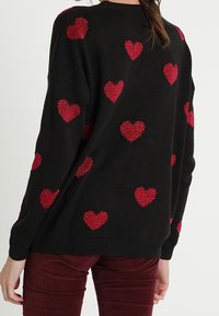 Musta neulepaita, jossa on punaisilla helmillä koristeltuja sydänkuvioita; rento istuvuus, resorireunuksilla kauluksessa ja hihansuuissa, pehmeä tekstuuri.