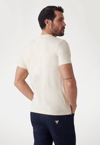 Camiseta de manga corta en color crema, hecha de una tela suave, con un diseño ajustado. Se lleva con jeans azul oscuro, mostrando un logotipo triangular en el bolsillo.