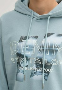 Hellblaues Kapuzenshirt aus weichem Stoff, mit einem auffälligen Grafiken "DENIM TOM TAILOR" und einem Landschaftsbild. Die Kordeln haben metallische Spitzen.