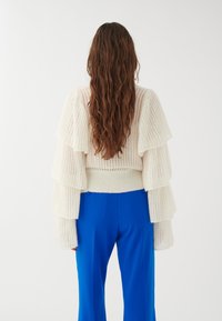 Maglione bianco a coste con maniche a volant, abbinato a pantaloni blue a gamba larga. Il maglione ha una finitura morbida e strutturata.