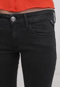 Jeans de mezclilla negros con un diseño ajustado, que cuentan con un botón plateado, bolsillos laterales y una pequeña etiqueta de la marca en la cinturilla.