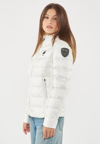 Giacca a vento bianca con cuciture orizzontali, colletto alto, tasche con zip e un patch nero con logo sulla manica sinistra. Texture liscia.
