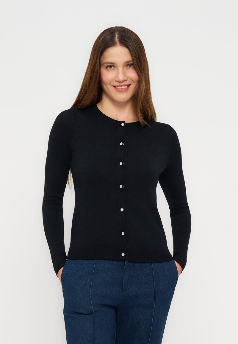 Γυναίκα με μακριά καστανά μαλλιά φοράει εφαρμοστό μαύρο κασμιρένιο πουλόβερ με κουμπιά και μπλε παντελόνι, στέκεται με τα χέρια στις τσέπες και χαμογελάει.