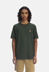 Camiseta de algodón verde con mangas cortas, con ribete dorado en el cuello y los dobladillos, que presenta un pequeño logo bordado en el lado izquierdo del pecho.