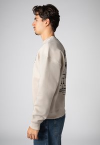 Beige sweatshirt with a round neck and ribbed cuffs, featuring black graphic print on the back. Model wears blue denim jeans.
