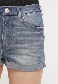 Džínové šortky v jasně modrém odstínu, s vysokým pasem, ohrnutými lemy a klasickým pětibokým designem. Viditelné stříbrné zapínání na knoflík.