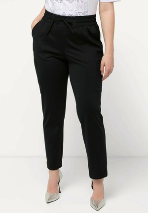 Vrouw die zwarte cargo-broek met hoge taille en zakken draagt, gecombineerd met zilverkleurige puntige hoge hakken, staand tegen een witte achtergrond.