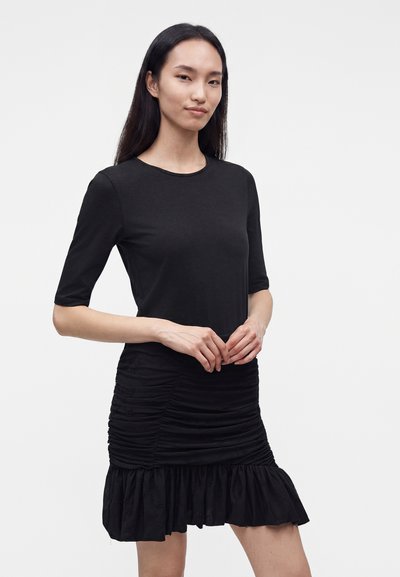 Femme aux longs cheveux foncés portant une robe noire ajustée à manches mi-longues, jupe froncée et ourlet à volants, debout devant un fond uni.