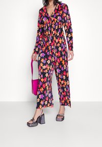 Kombinezon w kwiaty z długimi rękawami, ozdobiony wyrazistymi czerwonymi, purpurowymi i żółtymi kwiatami na czarnym tle. Do tego błyszczące buty na platformie i różowa torebka.