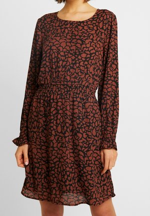 Kobieta w brązowej sukience z długim rękawem, ozdobionej czarnym abstrakcyjnym kwiatowym wzorem i elastyczną talią, stojąca na jednolitym jasnym tle.