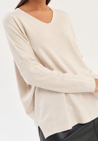 Beige v-ringad tröja med lös passform och sänkta axlar. Ribbad nederkant och ärmslut, tillverkad av mjukt stickat material. Slät textur.