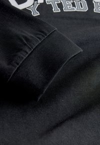 Sort langærmet beklædningsgenstand med tekstureret stof i close-up, med ribstrikket manchet, der viser diskret syningsdetalje.