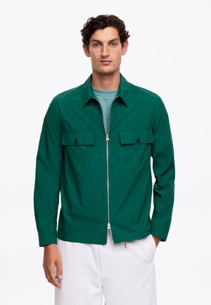 Uomo con giacca verde scuro con cerniera e due tasche sul petto sopra una maglietta turchese e pantaloni bianchi, in piedi davanti a uno sfondo neutro.