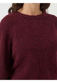 Femme portant un pull duveteux bordeaux profond avec un col arrondi et des manches longues, visible des épaules à la taille.