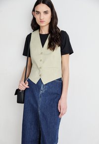 Gilet vert clair au design ajusté, cinq boutons et deux poches avant, associé à un t-shirt noir et une jupe en denim avec une fente à l'avant.