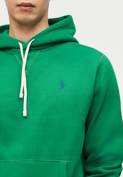 Moški v zeleni kapucarji z kremnimi vrvico za zategovanje in majhno modro vezeno značko na levi strani prsnega koša, beli ozadje.