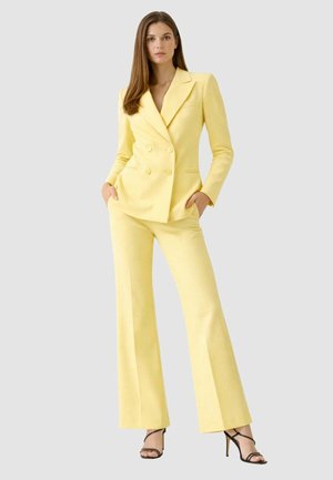 Donna in piedi con le mani nelle tasche, indossa un blazer doppiopetto giallo pallido su misura e pantaloni a gamba larga abbinati con sandali neri con tacco.