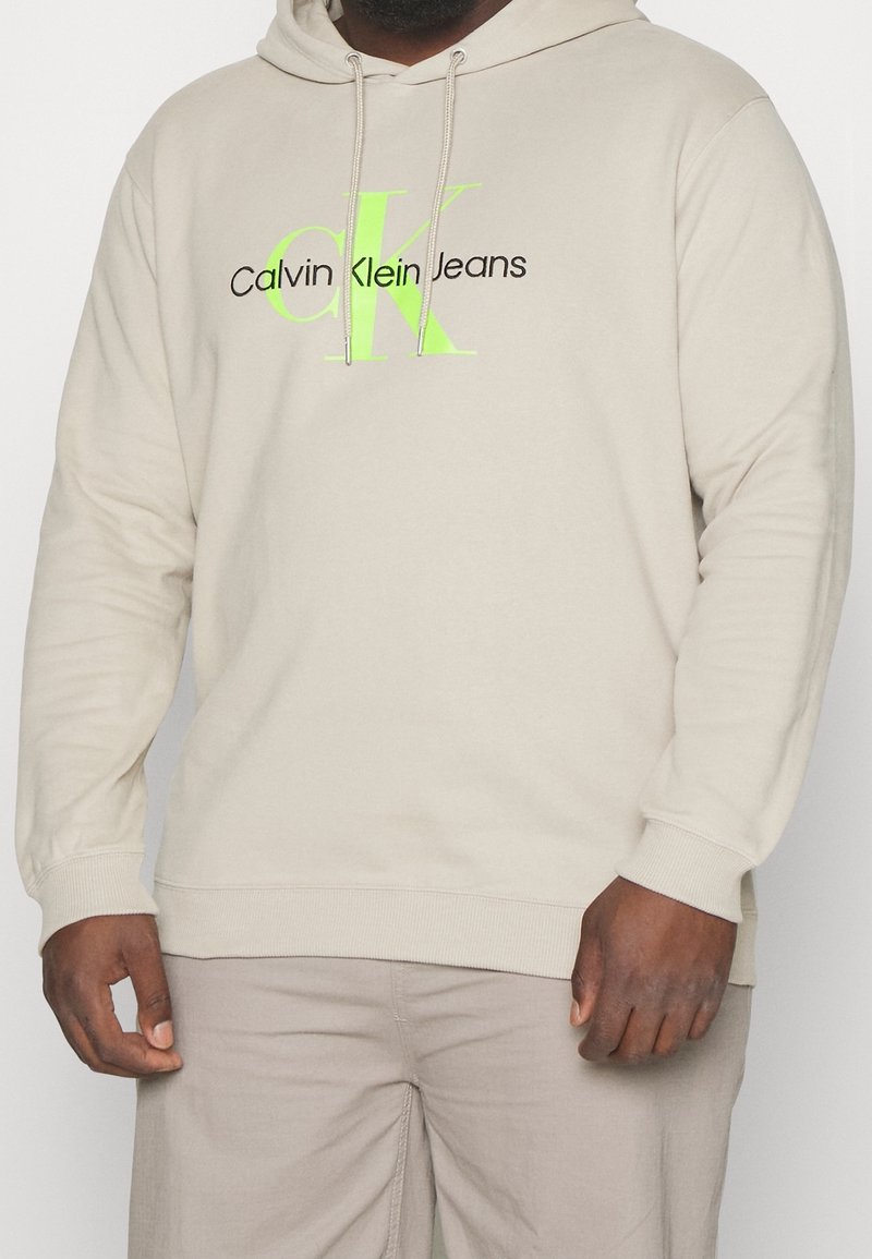 Homme portant un sweat à capuche beige Calvin Klein Jeans avec un logo "CK" vert néon et du texte noir, assorti à un pantalon beige clair.