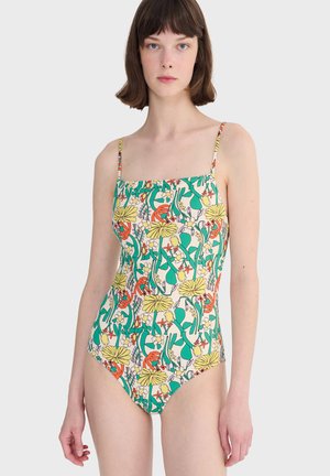 Ung kvinde iført en ærmeløs heldragt badedragt med et farverigt blomster- og bladmotiv på en lys baggrund.