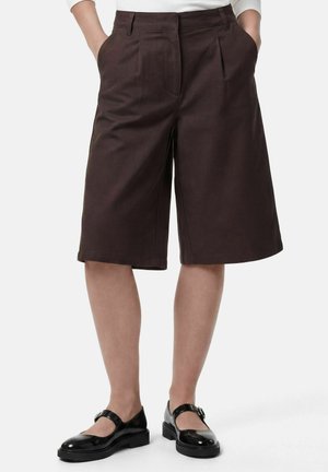 Short marron sur mesure arrivant aux genoux, porté avec des chaussures Mary Jane noires et un haut blanc, mains dans les poches, sur un fond uni.