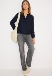Blouse navy à manches longues avec un décolleté en V et patte de boutonnage, associée à un jean droit gris clair et des baskets beiges. Sac à épaule crème.