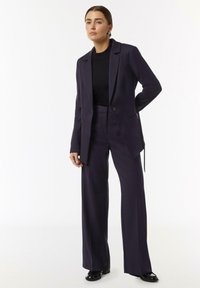 Granatowy garnitur z dopasowaną marynarką na jeden guzik i spodniami o szerokich nogawkach, w połączeniu z dopasowanym czarnym swetrem i czarnymi butami.