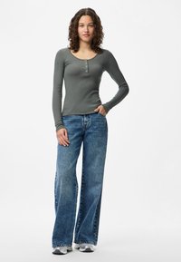 Donna con capelli ricci che indossa un top grigio aderente a maniche lunghe, jeans blu a gamba larga e sneakers bianche, in piedi con una mano in tasca.