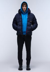 Marineblauwe pufferjack met een capuchon, voorzien van een rode voering en logodetail. Draag het over een felblauwe hoodie en zwarte broek, gecombineerd met zwarte schoenen.