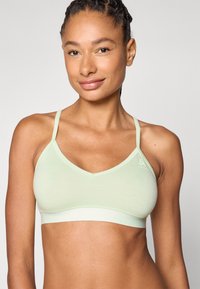 Bralette verde claro con diseño de escote en V, tirantes finos y una textura de algodón suave. Presenta un logo en el lado izquierdo y una banda elástica.