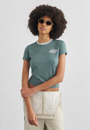 Γυναίκα με αφροχτένισμα που φοράει οβάλ γυαλιά ηλίου, πράσινο κοντομάνικο μπλουζάκι, ασημένιο κολιέ αλυσίδα και μπεζ ριγέ παντελόνι σε απλό φόντο.