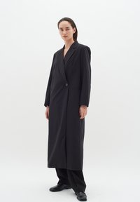Ilgas juodas paltas su struktūruota siluetu, turintis įpjautą apykaklę, vieną sagą, ir šonines kišenes. Lygus audinio paviršius.