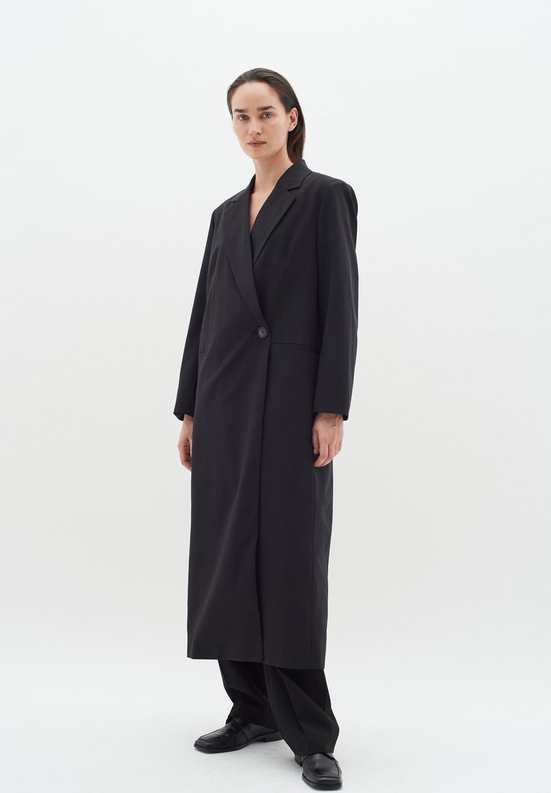 Ilgas juodas paltas su struktūruota siluetu, turintis įpjautą apykaklę, vieną sagą, ir šonines kišenes. Lygus audinio paviršius.