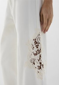 Pantaloni bianchi in tessuto con un'apertura laterale rifinita da pizzo floreale. Design a gamba dritta con superficie tessurizzata. Mano appoggiata sul tessuto.