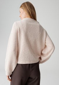 Maglione rosa chiaro con trama, vestibilità ampia e maniche lunghe, abbinato a pantaloni marrone scuro. Vista posteriore che mostra il morbido tessuto a maglia.